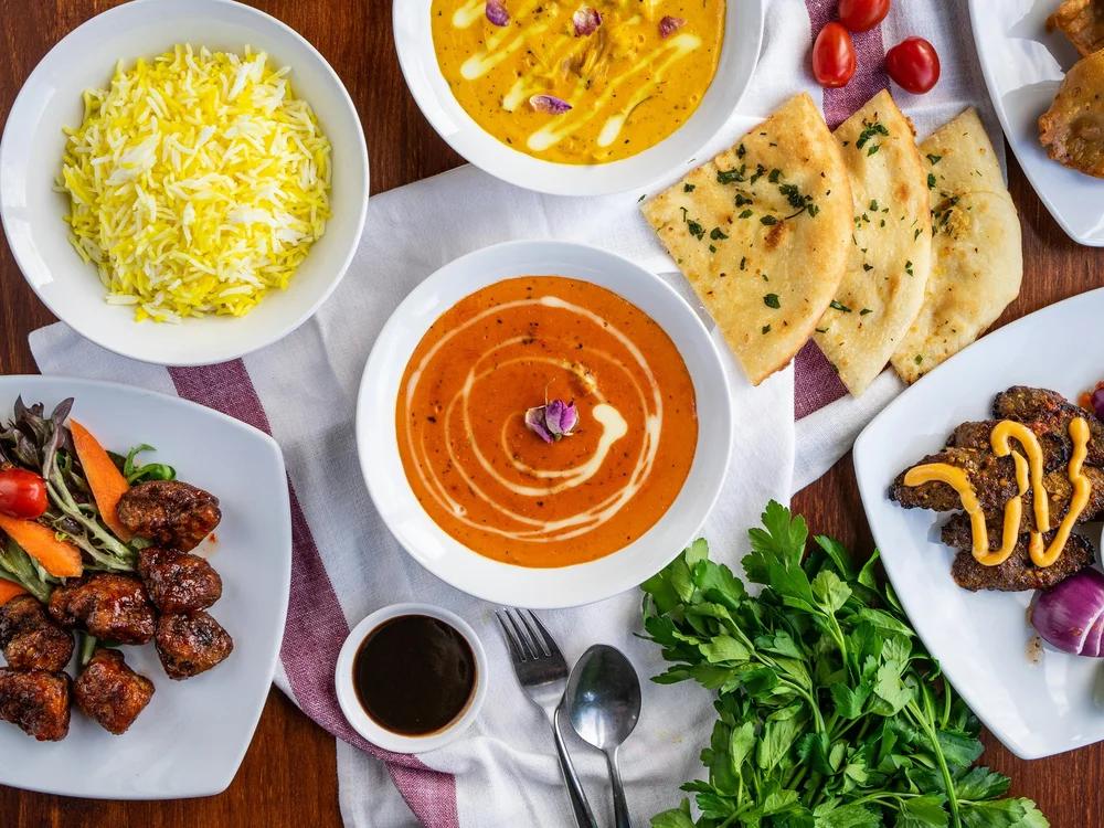 A variety of Indian dishes, including rice, curry, naan bread, and fried appetizers on white plates, with fresh herbs and cherry tomatoes decoratively arranged on a table.